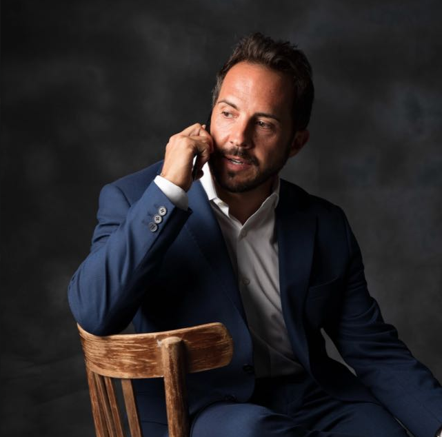 Uomo in abito blu navy seduto su una sedia di legno mentre parla al telefono su sfondo scuro.